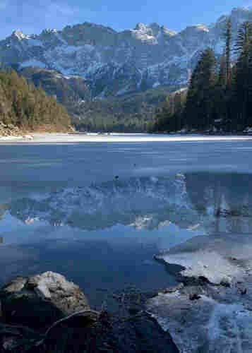 An alpine lake, partly frozen, with rocky mountains in the background, which reflect on the water in the foreground.