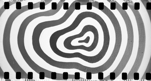 A black and white photograph taken with a film camera, showing the film sprocket holes along the top and bottom edges. The image features a mural with concentric wavy lines forming a pattern resembling a topographical map. The lines vary in thickness and alternate between light and dark shades, creating a hypnotic visual effect.
