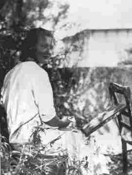 Charlotte Solomon - a young woman sitting in the grass. In front of her a canvas for painting a picture and a chair. Her head is turned slightly to the right. A blurred tree and building is visible in the background.