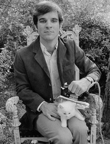 B&W photo of Steve Martin plays it straight, while posing in a chair and ironing a fluffy white kitten.