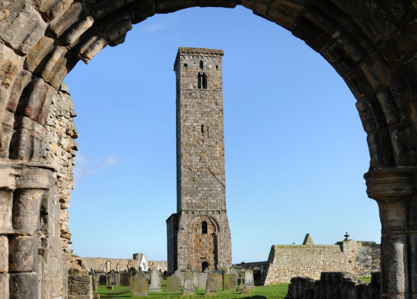 St Rule's Tower in St Andrews. The image shows a decorated but very weathered stone arched doorway surrounding the frame. Through it can be seen a very tall and narrow stone tower, surrounded by gravestones. There are stone walls on the right and in the background, The sky is blue.