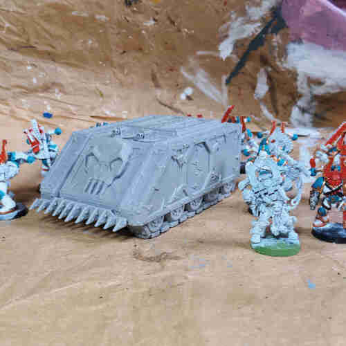 An unpainted light grey model tank with a large skull on the front and a spikey ram surrounded by old games workshop models of khorne berserkers that are half painted. The background is brown paper stained with various colours of paint.