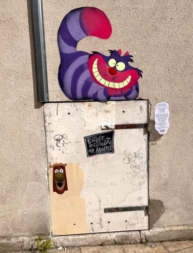 Streetartwall. A purple Cheshire cat has been painted on a beige-colored street wall above a small metal door. It has a red and purple face and tail and a purple striped body. She is lying there, grinning and looking directly at the viewer. On the door it says "Entry forbidden to adults" in French. The doorknob is adorned with a friendly face with an open book cover as a hairstyle, also a character from Alice in Wonderland. Next to the mural is a note with a handwritten text.
Info: Alice in Wonderland is a children's book by Lewis Caroll. In it, the Cheshire Cat represents the absurd and mysterious world into which Alice plunges. The small handwritten text next to the mural is in French and means something like: Preserve the imagination of childhood, then everything is possible and life can be a celebration.