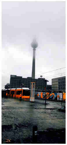 Foto vom Fernsehturm in Berlin, Alexanderplatz. Die Spitze des Turms verschwindet im Nebel. Die orangenen Farbtöne einer im Vordergrund haltenden Tram und das orange e Graffiti an einem Holzzaun um eine Baustelle, sind extra hervorgehoben.