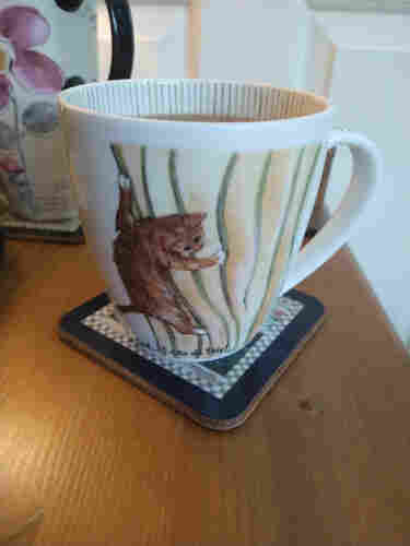 Mug of tea on a bedside cabinet, the image shows a ginger kitten clinging to green and yellow striped curtains, underneath it says 'And.... I can do this'