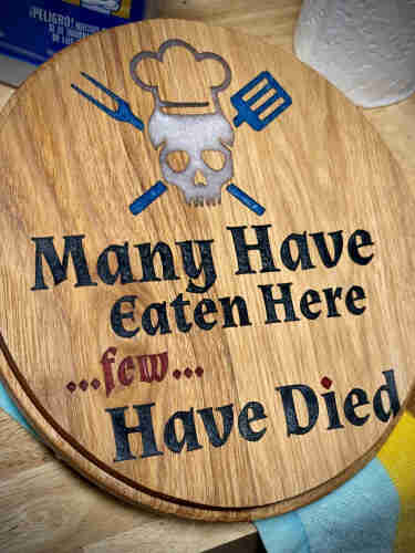 A wooden cutting board with the phrase "Many Have Eaten Here... few... Have Died" and a graphic of a skull wearing a chef's hat crossed by a fork and a spatula.