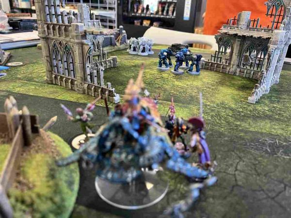 Warhammer 40K Battle

A unit of brightly colored harlequins jump out of a brightly colored star weaver to claim an objective. 

Space marine intercessors approach from the other side of some ruins, stern guard follow behind them. 