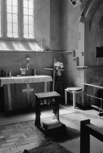 Portrait format black and white photo showing light falling through a clear church window onto an altar and a prie-dieux kneeler.
