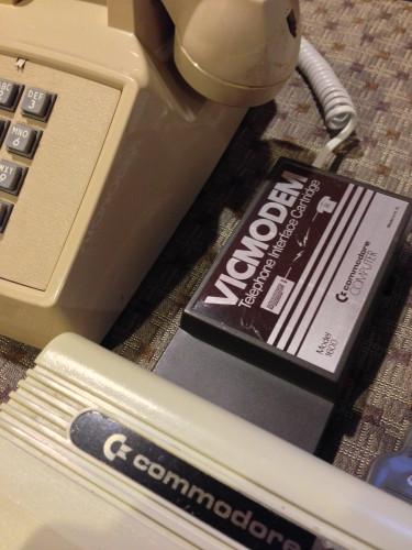 Photo of the right corner of push button phone, the top left corner of a Commodore VIC-20 at the bottom, and a brown and silver VICMODEM cartridge plugged into the back of it. A telephone coil cable was removed from the handset and plugged into the modem.