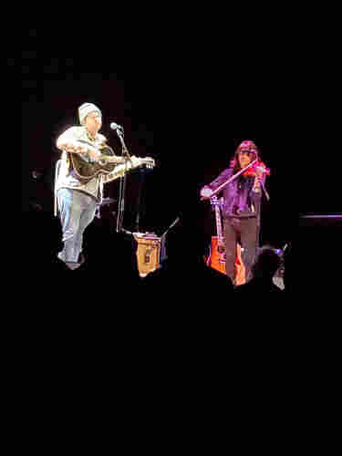 Singer songwriter Abe Partridge (left), wearing jeans, plaid shirt, toque, playing guitar, performs with violinist from Matthew Sweet's band (right), with concert attendees silhouettes below them