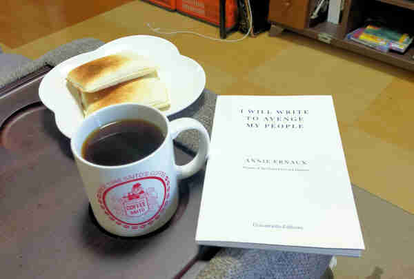 The photo is of a brown couch divider, but the couch is gray. Nestled in the cup holder is a white coffee mug of black coffee with the red medieval-esque logo of Saito's Coffee. To the right is the small white paperback book with blue lettering. Beyond the mug is a plate of 2 toasted square bread.