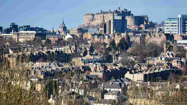 A photo of a landscape, a city on a hill fills the frame. There are many buildings, mostly of brown or grey brick, but a few are stone-built and a few kirk spires can be seen. Only a few modern structures are in the scene. Centre right, atop a hill, is an imposing castle rising above all else in the shot.