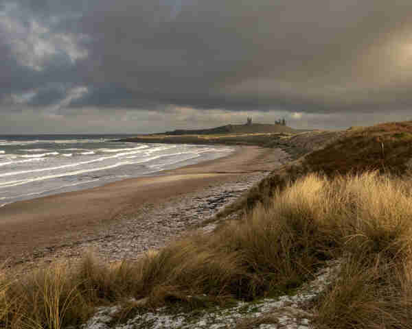 A sandy beach with dunes behind curves out towards the sea. Behind the dunes in the distance is the ruin of a castle. The sky is cloudy with stormy clouds above the castle. In the foreground the tops of dune grasses are catch the sun as it breaks through the clouds to the right of the picture..