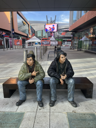 A statue of beloved SCTV characters Bob and Doug Mackenzie, in toques and parkas, holding brown stubby beer bottles sits at the entrance of the south entrance to Rogers Place. Behind them on a giant video screen we see a shot of Oilers' captain Connor McDavid celebrating a goal.
