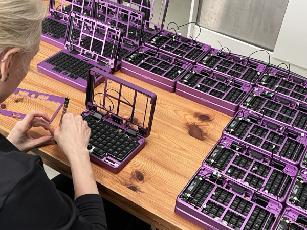 greta working with a stack of magnets on a purple half assembled mini laptop. there afe many of those laptops on a wooden table