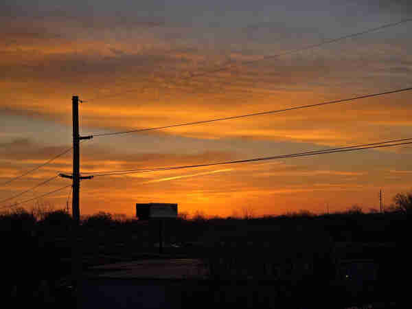 The sunrise over St. Louis, Missouri this morning. There's a very nice blast of oranges with bands of blue. Unfortunately, there are also power lines and poles and a billboard, it's an industrial park. Dark trees line the bottom.