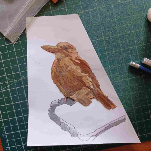 A work in progress veneer marquetry image of a kingfisher sitting on a branch, looking to the left, and rendered in various shades of orange, red-brown, brown, peach & beige. Still to be made: the feet, the branch it is sitting on, and the background.