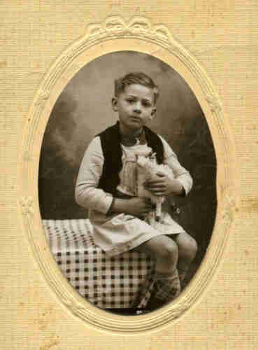 Black and white photograph in an oval pass-partou. A boy sits on a bench covered with a checkered tablecloth. He is wearing a white long blouse and a black waistcoat. In his hands he trisma a toy sheep on wheels.