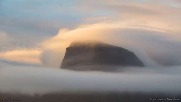 A photo of an oddly shaped mountain, with a wide, flat, near-vertical front and sloping sides. The dark rock is mostly hidden by wreaths and scarves of cloud, one layer is quite low and obscures part of the lower slopes completely. Above the mountain top is a lovely, curved cap of cloud, picking up the hues of lowering sunlight. Distinct layers are visible in this disc-shaped hat.