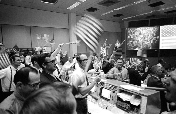 black and white photo of a room of jubilant nasa engineers waving flags and looking really excited