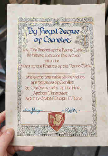 Parchment paper with a certificate of Knighthood calligraphied onto it, and decorated with a black ink and metallic gold border of swirling vines, leaves, and flowers.

At the top in big blue ink:

'By Royal Decree of Camelot'

Beneath, in smaller brown ink;

'We, the Knights of the Round Table, do hereby welcome the acclaim into the Order of the Knights of the Round Table (space for someone's name) and grant said noble all the rights and privileges of Camelot by the divine right of the King, Arthur Pendragon, and the Grand Wizard Merlin"

There are two signatures on the left and right at the bottom; Arthur Pendragon in swirly cursive, and Merlin with two stars either side, in curly uncial script, and the 'i' dotted with a heart.

Beneath that, the crest of Camelot - a dark red shield with a gold dragon and a small triskelle under it.