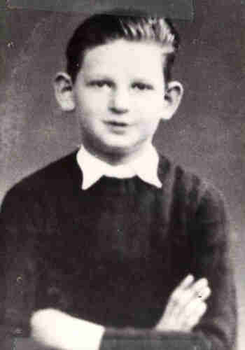 Black and white portrait photograph of a young boy. He is photographed from the waist up. His arms are folded. He is wearing a black jumper with a white collar sticking out from underneath. His short hair is combed up and his ears stand out slightly. 