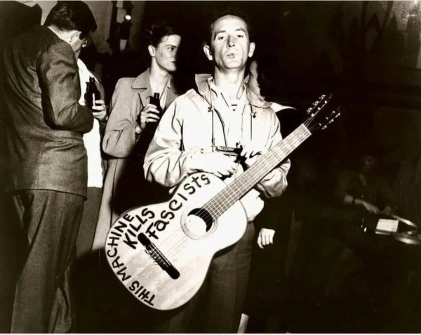 image alt: guthrie with guitar that says "This Machine Kills Fascists" in front of a few people kicking it in formal attire with dark bottles.