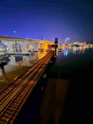 Colorful night view overlooking the Florida East Coast Railway tracks and drawbridge trestle crossing the Saint Johns River.  The rusty old tracks shine in orange and red from nearby illumination. Adjacent is the Acosta traffic and pedestrian bridge, massive grey concrete structures brightly painted with cow murals and illuminated with bright yellow lighting.  The far shoreline of the calm, wide, dark river is lit with the colorful night lights from a skyline of buildings all casting reflections upon the still waters below., all below a clear deep blue night sky.