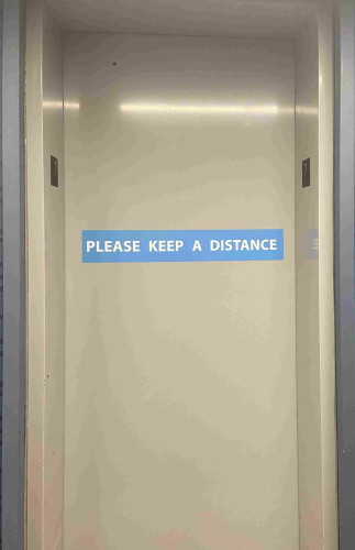 Photo of an elevator door with a large sign reading “Please keep a distance.”