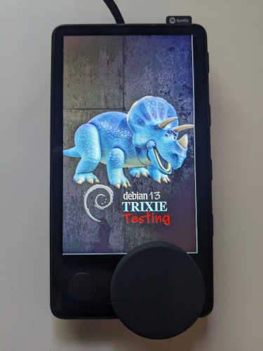 Photo of the (formerly known as) Spotify Car Thing. It's a small, thin black, phone-looking device that is mostly screen, a fairly thin bezel, small button in the lower-left corner, and a large knob partially hanging off the bottom right corner.
The screen is displaying a boot logo of a blue toy triceratops and the text:
"debian 13
TRIXIE
Testing"