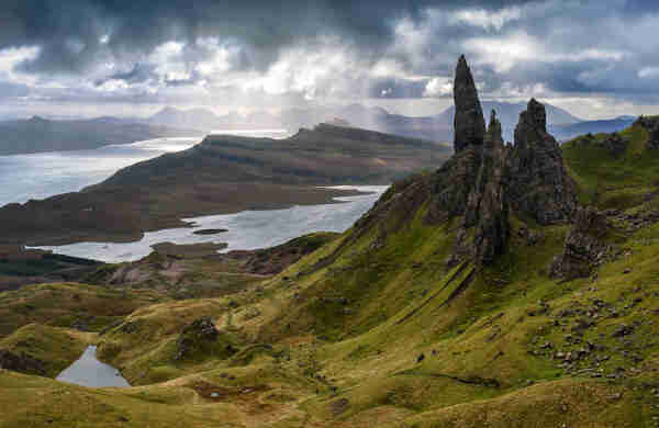 A view of Storr, Scotland, with craggy mountain peaks looming above green hills and fjords below. In the distance, sunbeams slant down through gaps in the cloudy sky.