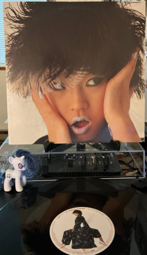 A record sleeve on a stand over a turntable. Sleeve portrays a woman with feathered hair and light blue lipstick with her hands to her face.

Below is a record on the turntable. The center label shows the same woman with  her legs on either side of a box and looking over her shoulder.