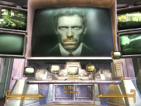 Still image. Screen capture from Fallout: New Vegas (2010). An enormous screen flanked by four smaller ones above a bench with several table-top computers. The screen is dominated by the face of Hugh Laurie in green tones. 

The cursor text reads:
E) Talk
Mr. House