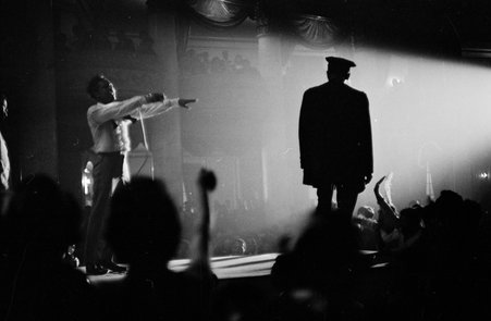 Blurry image of Jackie Wilson on stage in a dark theatre with his arms outstretched. The shadowy figure of a policeman or security guard can also be seen standing on the stage.