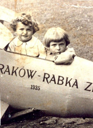 Two children are seated inside the cockpit of an old-fashioned airplane with the text "Kraków-Rabka Zdrój 1935" painted on its side. The child on the left is looking at the camera with curly hair, while the child on the right has straight hair and a serious expression, looking forward.