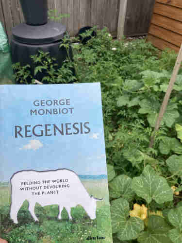 The book Regenesis by George Monbiot. Held up in front of a vege patch sprawling with tomatoes and pumpkin and a black compost bin behind
