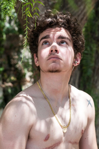 Photo of a shirtless trans man with dark curly hair standing in a sunny spot with trees behind him, looking upwards