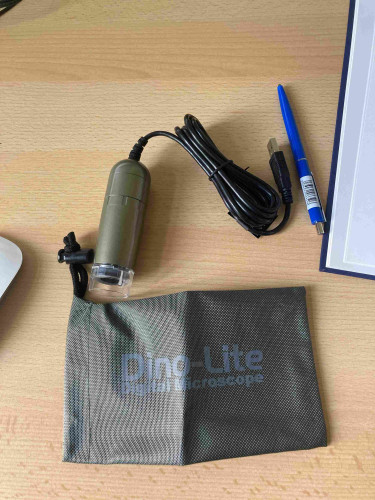 A digital microscope with a USB cable and a storage pouch labeled "Dino-Lite Portable Microscope," next to a blue pen on a wooden desk.