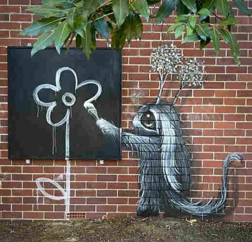 Streetartwall. A mural with a fluffy fantasy figure was sprayed/painted on a brown brick wall in a park. A small gray-black, fluffy creature with big eyes, curved ears, a broad nose and two tufts of plants on its 2head stands there with a paintbrush in its hand. It looks like a mixture of squirrel, mouse and rabbit. It is drawing a white flower, like a child's drawing, on a black board and the stem on the wall below. (Green leaves hang into the picture from the top right.
Info: The mural is part of a larger picture on this wall. There are two more fluffy creatures on the right-hand side.(Extra-Photo Part 2/2)