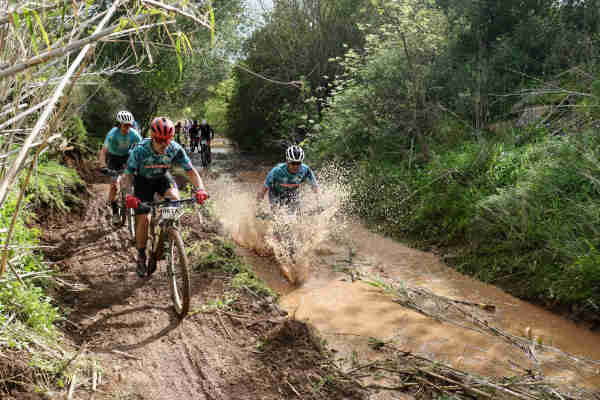 Mountain bikers riding through a muddy water crossing in a forested area.