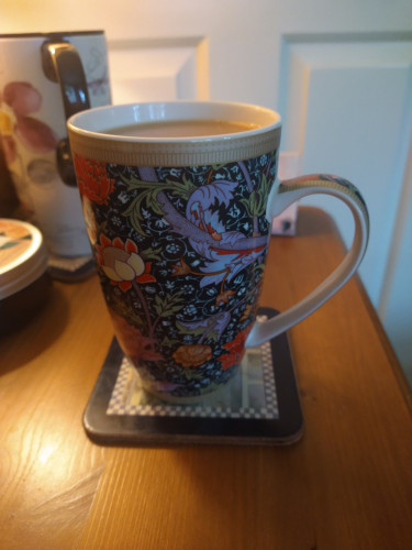 William Morris mug of tea on a bedside cabinet.
It has a dark blue background and is overlaid with a gold, pink, purple and red floral design 