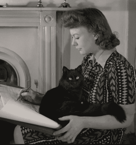 Black and white photo of a young white woman is badly patterned dress, working at a drawing table. In her lap is curled a gorgeous longhaired black cat that looks brightly up at the camera.