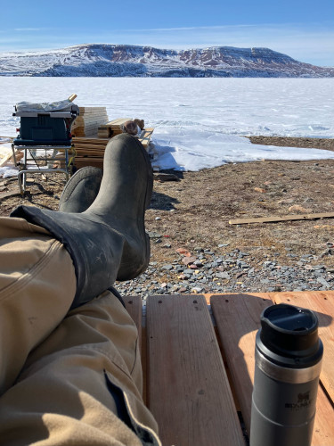 Legs of a man sitting on a deck, brown pants and green rubber boots. Beside him on the deck is a thermal coffee mug. In front of him is a table saw and a pile of money. Beyond that is a frozen ocean and across that mountains. 