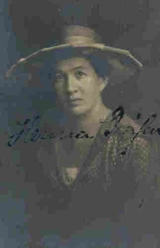 Portrait photograph of a mature woman. She is wearing a hat with a very wide brim on her head. She is wearing a blouse with a dotted pattern. Around her neck is a chain with a pendant.