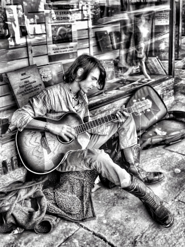 A busker sitting on the sidewalk in front of a charity playing his guitar in black and white in Totnes, England