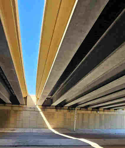 Beneath a double overpass bridge looking upwards at the space between where a brilliant and sunny clear blue sky casts an angled light beam into the shadows below, surrounded by the old weathered concrete bridge structure.