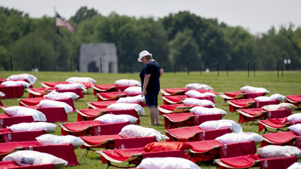 A person stands amid rows of cots on a grassy lawn, part of the 2023 Millions Missing demonstration for patients who have Long Covid and ME/CFS (myalgic encephalomyelitis/chronic fatigue syndrome). Each cot has a pillow with a pillowcase that has been decorated by a patient.