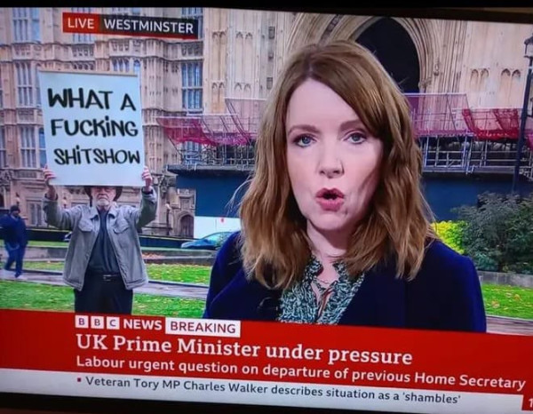 Bbc news. Woman reporting from Westminster. Ticker reads UK Prime Minister under pressure. In the background a man stands with a handwritten sign that reads WHAT A FUCKING SHITSHOW. 