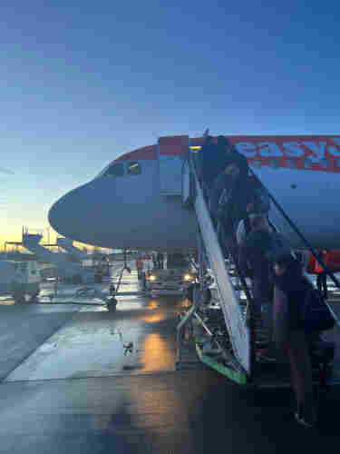 The front of an easyJet aeroplane, with people boarding up a flight of metal steps. It is early in the morning, and just after dawn. 