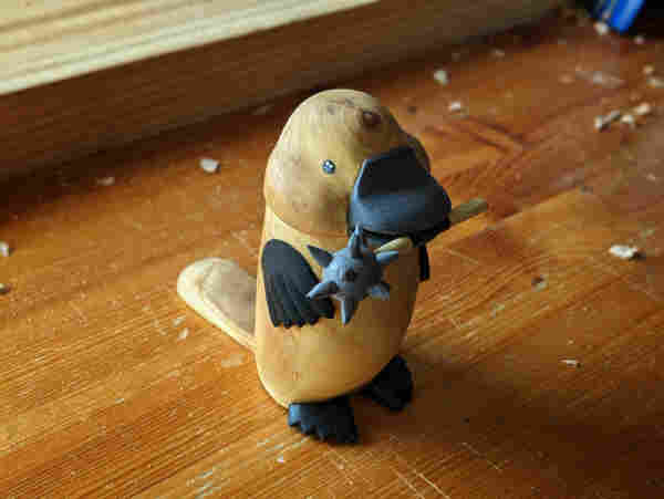 Woodcarved standing platypus holding a morningstar in its beak